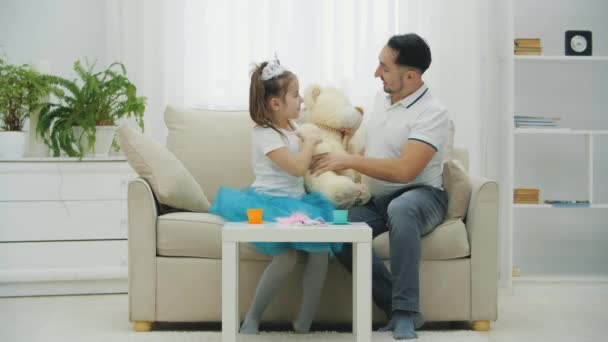Nettes Mädchen und ihr Vater sitzen auf dem Sofa und umarmen Teddybär. Vater rührt Töchtern Nase an, dann Teddys. — Stockvideo