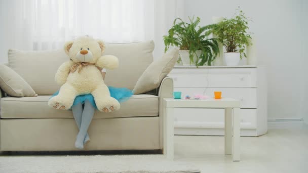 Pequena princesa em coroa e vestido está sentado no sofá, escondido atrás do ursinho de pelúcia . — Vídeo de Stock