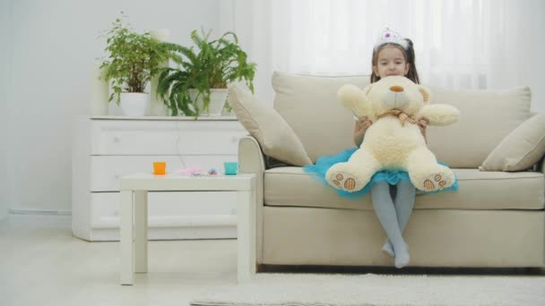 Pequeña princesa en la corona y el vestido está sentado en el sofá, sosteniendo oso de peluche en sus rodillas. — Vídeos de Stock