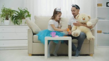 Baba, kız ve Teddy çay partisi veriyorlar. Kanepede oturuyorlar, çay içiyorlar. Komik baba ve taçlı tatlı kız..