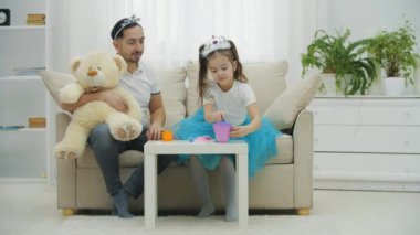 Küçük prenses çaydanlıktan renkli bardaklara çay dolduruyor. Baba, kız ve ayıcık çay partisi veriyor..
