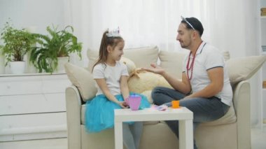 Baba ve kızı prenseslerle oynuyor. Kızların elini öpüp ısırıyormuş gibi yapan komik bir adam. Ona sarılıyor ve bir taç kırıyor. Bebeklerin gözleri yaşarır.