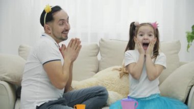 Komik baba ve küçük kızı kanepede oyuncak taçlarla iğneleyerek kraliçelerin zarif tavırlarını taklit ediyorlar, aptal gibi görünüyorlar..