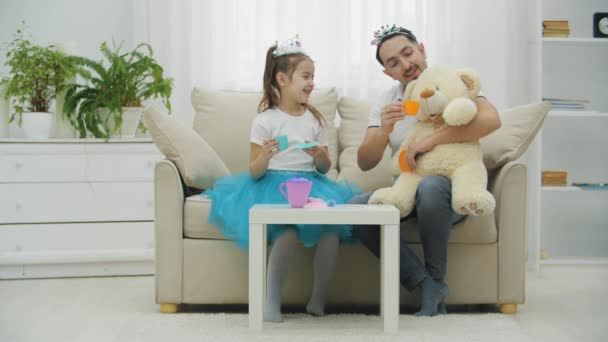Vader, dochter en teddy houden theekransje. Ze zitten op de bank. Grappige vader en schattig meisje in kronen. — Stockvideo