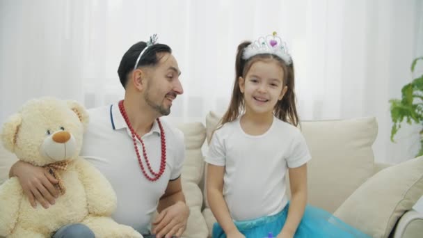 Père joyeux joue avec sa fille. Ils sont assis sur le canapé en couronnes princesse, étreignant, tenant ours en peluche, souriant. — Video