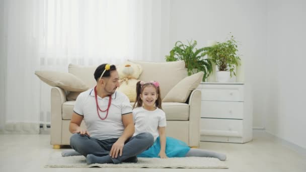Playful father sitting on the floor in lotos position, his little daughter making split. Ther are in princess crowns, dancing, moving hands and hips comically. — Stock Video