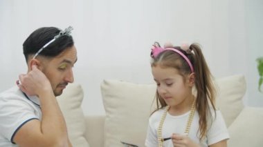 Küçük çocuk babasına makyaj yapıyor, göz kapaklarına yeşil gölgeler koyuyor. Babam aptal bir ifadeyle kameraya bakıyor..