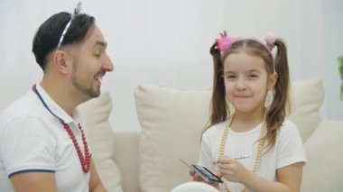 Küçük çocuk babasına makyaj yapıyor, göz kapaklarına yeşil gölgeler koyuyor. Babam aptal bir ifadeyle kameraya bakıyor..