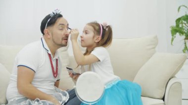 Sevimli küçük çocuk babasına makyaj yapıyor, göz kapaklarına sarı ve menekşe rengi gölgeler sürüyor..