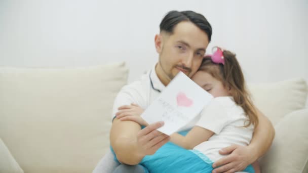 Unscharfe Nahaufnahme eines lustigen Vaters, der auf dem Sofa sitzt, seine kleine verschlafene Tochter auf den Knien hält, sie einlullt und ein Blatt Papier zeigt, auf dem der Tag des glücklichen Vaters steht. — Stockvideo