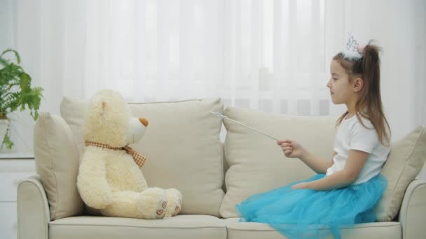 Kleine Fee sitzt auf dem Sofa mit Zauberstab in der Hand, zaubert ihren Teddybär herbei und richtet dann den Zauberstab in die Kamera. — Stockvideo