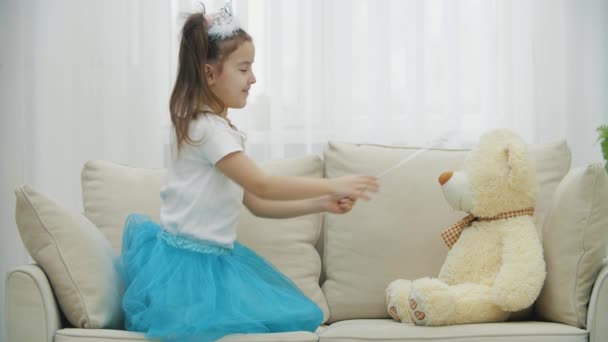 Kleine Fee sitzt auf dem Sofa mit Zauberstab in der Hand, zaubert ihren Teddybär herbei, richtet dann den Zauberstab in die Kamera und lächelt. — Stockvideo