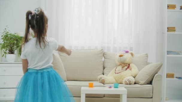Blurred little fairy standing on the forefront with magic wand in her hand, conjuring her teddy bear on the sofa, then turning to the camera, smiling. — Stock Video