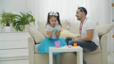 Komik baba ve kızı prenseslerle oynuyorlar, kanepede oturuyorlar, taçlar giyiyorlar. Babam küçük kızların elini öpüyor..