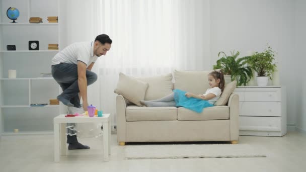 Petite fée et papa jouent, la fille est allongée sur le canapé, agitant la baguette magique, et papa obéit à toutes ses commandes. — Video