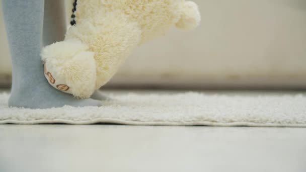 Cropped legs of little girl standing on white carpet, holding a plush teddy bear in necklace, putting its paw on her feet and teaching it how to dance. — Stock Video