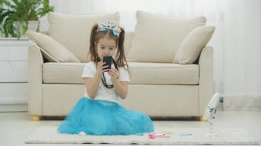 Taç takan, elbise takan, kolye takan ve elinde telefonla halının üzerinde oturan güzel bir çocuk..