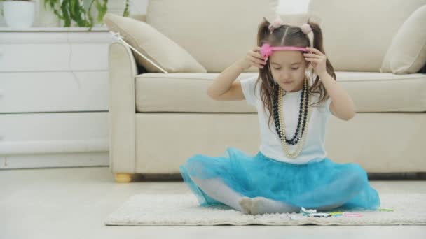 Ragazzina carina con le trecce, seduta sul tappeto, indossando copricapo, diverse clip di capelli e fiocchi. — Video Stock