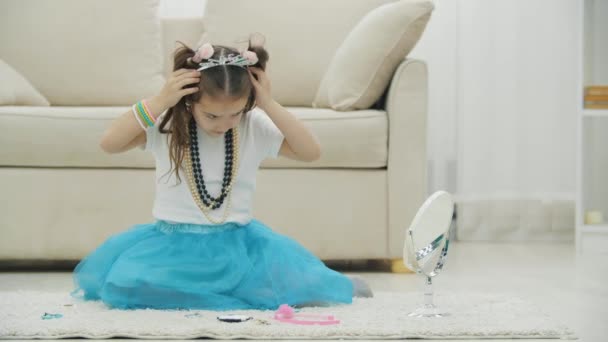 Nettes kleines Mädchen, das auf dem Teppich sitzt, in den Spiegel schaut, Armband, Halskette, Kopfbedeckung und verschiedene Haarspangen trägt, posiert. — Stockvideo