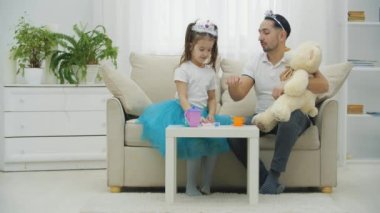 Tatlı prenses kız babasını güzelleştiriyor, kafasına taç takıyor ve eline de el bandı takıyor. Babam ayıyı dizlerinin üstünde tutuyor..