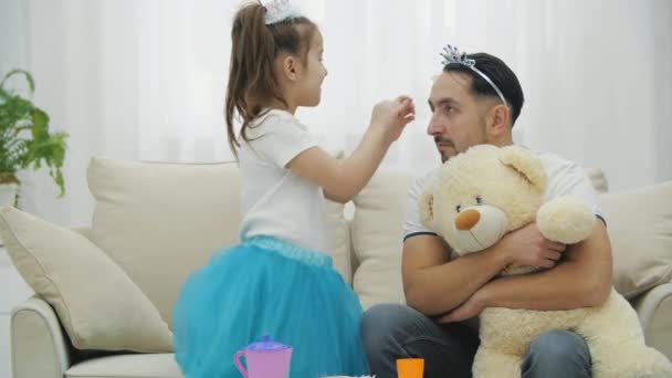 Dottern justerar kronan på fädernas huvud. Söt prinsessa flicka, hennes prinsessa pappa och Teddy har en tebjudning. — Stockvideo