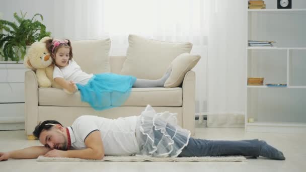 Roztomilá dívka mává kouzelnou hůlkou a tatínek v legračním oblečení poslouchá její příkazy, leží na podlaze, úplně unavený. — Stock video
