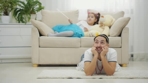 Lively father lying on the floor, his little daughter climbing on his back, they are giving thumbs up, smiling. — Stock Video