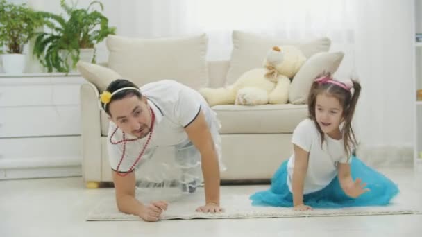 Jonge grappige vader en kleine schattige dochter doen oefeningen op het tapijt, gedragen in hoofddeksels. — Stockvideo