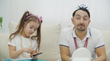 Şirin küçük kızı elinde fırça ve yüz pudrası tutuyor. Taclı ve kolyeli komik babası için makyaj yapıyor..