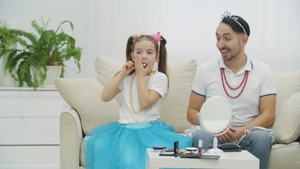 小女儿在玩化妆品刷的把戏.父亲坐在她身边，头戴项链，踌躇着. — 图库视频影像