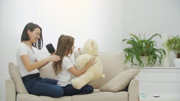 Ibu muda sedang menyisir rambut putrinya saat dia bermain dengan boneka beruang.. — Stok Video