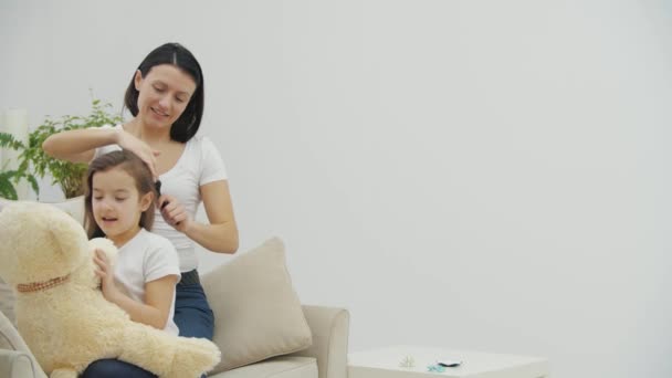 Jovem mãe está penteando o cabelo de suas filhas enquanto ela está brincando com ursinho de pelúcia . — Vídeo de Stock