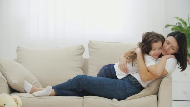 Linda niña y madre encantadora están tumbadas en el sofá, sonriendo, besándose y abrazándose. — Vídeo de stock
