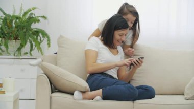 Genç anne elinde telefonla kanepede oturuyor, fotoğraflara bakıyor. Kanepenin arkasına yaslanmış uzun saçlı güzel kız çocuğu da ekrana bakıyor..