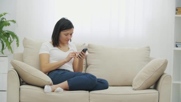 Charmig vacker kvinna sitter på soffan med mobiltelefon i händerna, surfa på internet eller meddelanden, benen hoprullade. — Stockvideo