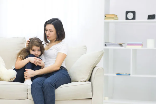 Foto de una niña triste sentada en el sofá y su mamá abrazándola. — Foto de Stock