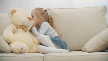 4K yavaş çekim videosunda küçük kız kanepede oturmuş, oyuncak ayısıyla konuşuyor ve sessiz işareti gösteriyor..