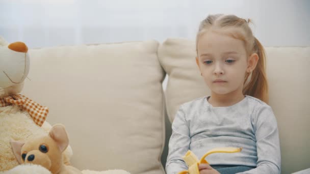 4k slow motion vídeo of little daughter eating yellow banana sitting on sofa. — Vídeo de Stock