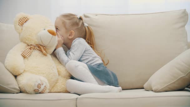 4k rallentatore video di bambina seduta sul divano, parlando con il suo orsacchiotto e mostrando segno silenzioso. — Video Stock