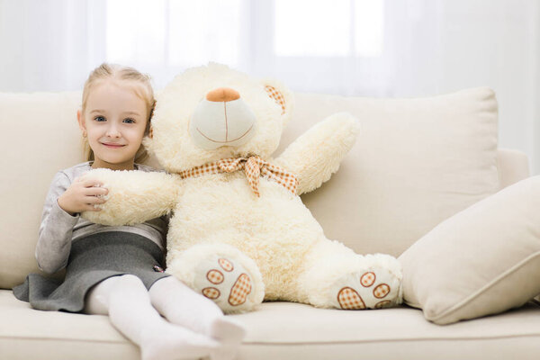 Cute little girl sitting on couch with teddy bear isolated on white background.