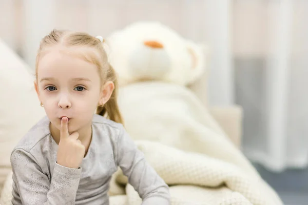 Menina bonito sentado com ursinho de pelúcia sob o cobertor. Menina está mostrando sinal de silêncio — Fotografia de Stock