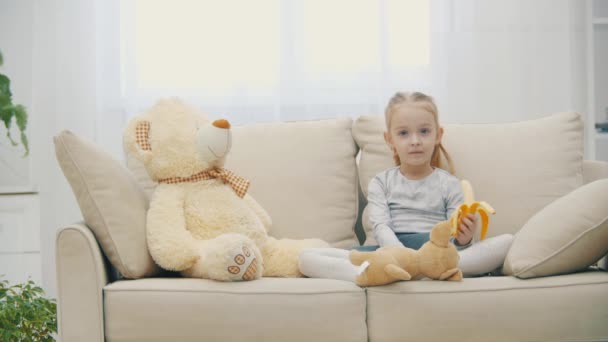 4k vídeo de pequena filha comendo banana no sofá e ursinho de pelúcia ao lado. — Vídeo de Stock