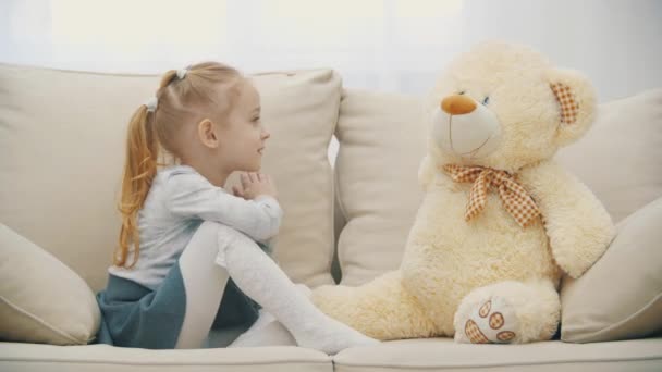 4k video de linda niña sentada en el sofá, hablando con su peluche. — Vídeos de Stock