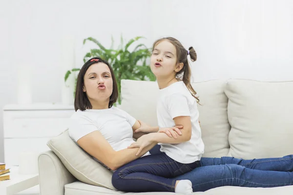 Foto de la madre y la hija sentadas en un sofá blanco y muestran un beso en la cámara. — Foto de Stock