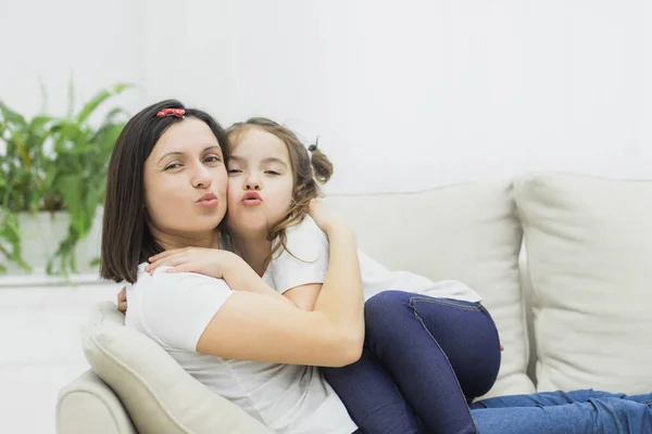 Foto de la madre y la hija sentadas en un sofá blanco y muestran un beso en la cámara. — Foto de Stock