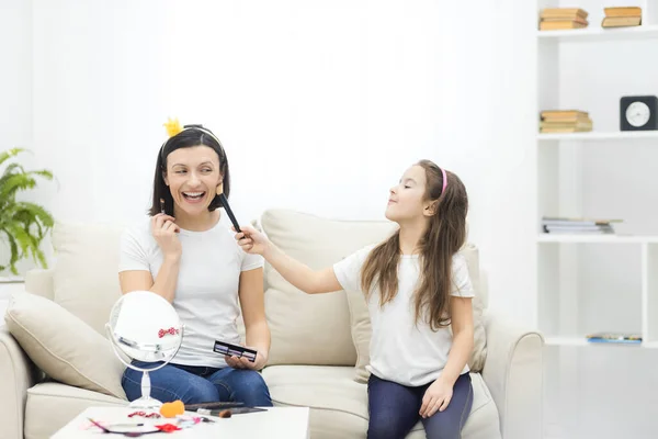 Junge hübsche Frau beim Make-up, während sie mit ihrer kleinen Tochter im Wohnzimmer sitzt. — Stockfoto