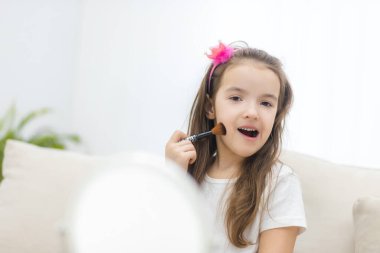 Kozmetik fırça tutan küçük tatlı bir kız fotoğrafı beyaz arka plandaki aynaya bakıyor..
