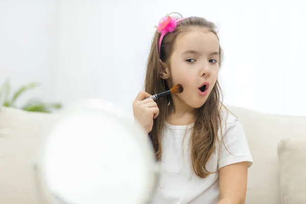 Photo de petite fille mignonne tenant pinceau cosmétique regardant le miroir sur fond blanc. — Photo