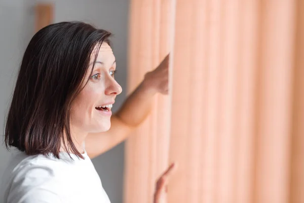 커튼이 달린 밝은 창을 바라보고 있는 행복 한 여인의 사진. — 스톡 사진
