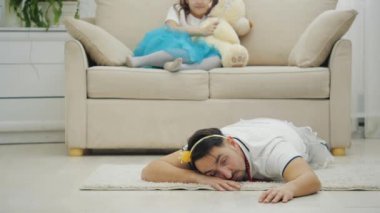 Tacın içindeki baba yerde bitkin bir şekilde yatıyor ve uyumak istiyor. Kızı enerji dolu, sihirli değnekle kanepede oturuyor ve oyuncak ayıyla oynuyor..